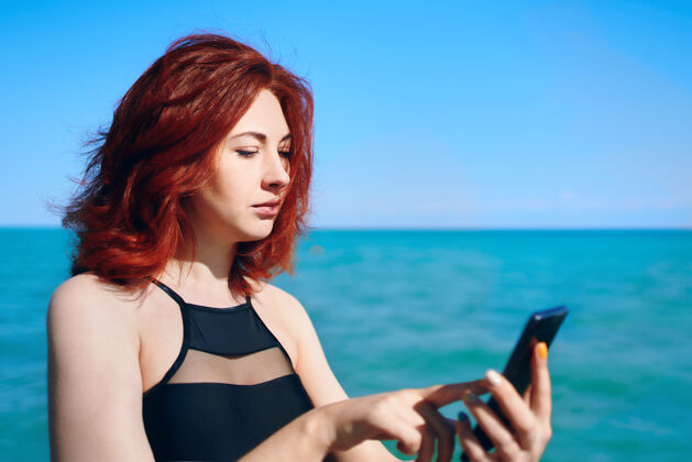 明亮红发女子对着大海用手机写短信海洋发送风景