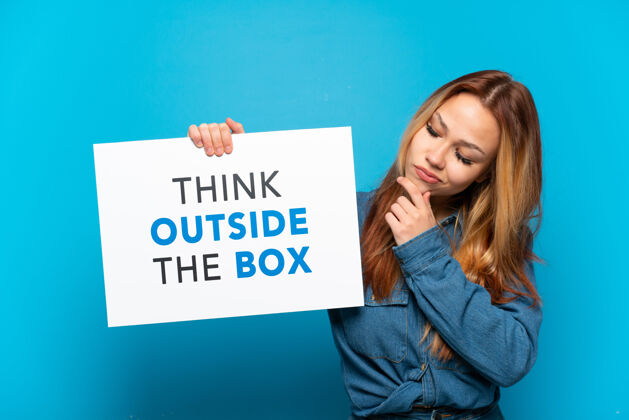 创新十几岁的女孩在孤立的蓝色背景下举着一个写着文字的标语牌 在盒子外思考纸板远见女人