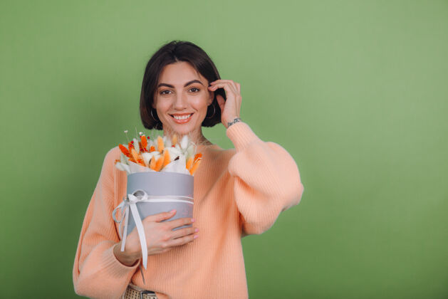 积极年轻女子穿着休闲桃色毛衣隔离在绿橄榄墙上手持橙白色花盒组成的棉花 吉普赛拉小麦和拉古鲁斯作为礼物开心惊喜内部黑麦美丽