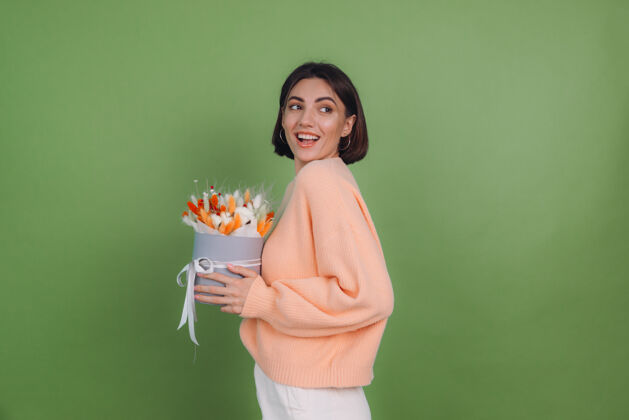 圆形盒年轻女子穿着休闲桃色毛衣隔离在绿橄榄墙上手持橙白色花盒组成的棉花 吉普赛拉小麦和拉古鲁斯作为礼物开心惊喜干花花束女性棉花花