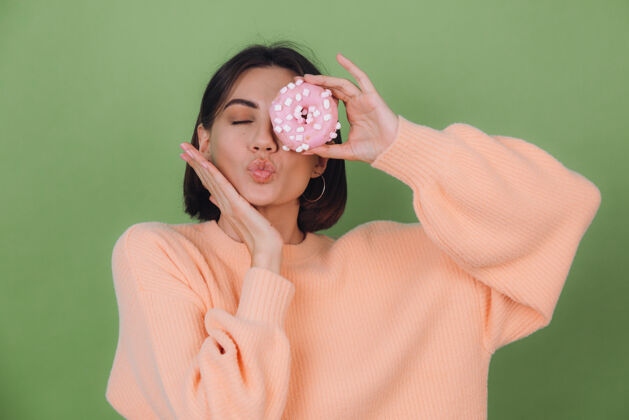模特年轻时尚的女士穿着休闲桃色毛衣隔离在绿色橄榄色的墙壁上送上亲吻与粉色的甜甜圈复制空间黑发脸情感