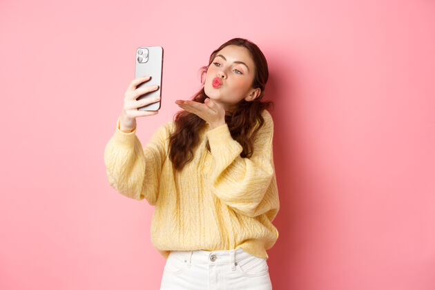 互联网漂亮的调情女孩在智能手机上自拍 视频聊天时送上空中接吻 站在粉红色的墙上复制空间快乐欢呼水平