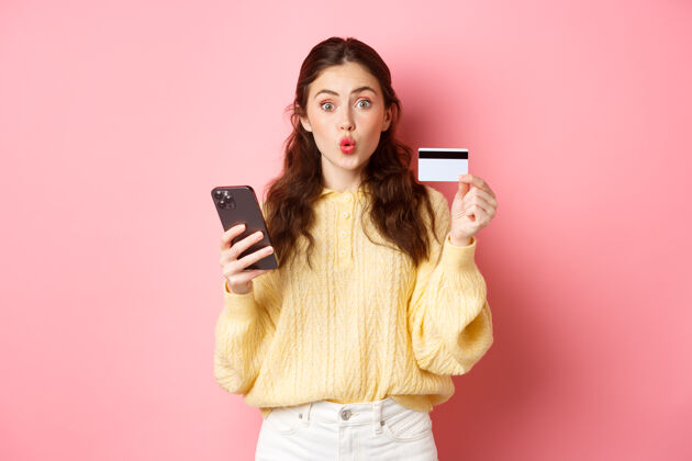 应用程序科技和网上购物兴奋的女孩购物者展示塑料信用卡 手持手机 用智能手机支付 在线订购 站在粉红色的墙上兴奋快乐聊天