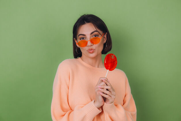 女孩年轻时尚的女士穿着休闲桃色毛衣 戴着橙色眼镜 隔离在绿色橄榄色的墙上 用橙色棒棒糖送上空气亲吻的复制空间青年青少年表情