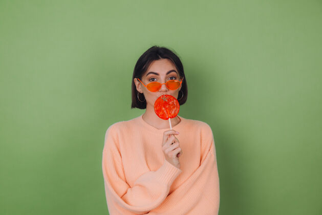 无忧无虑年轻时尚的女士穿着休闲桃色毛衣 戴着橙色眼镜 隔离在绿色橄榄色的墙上 用橙色棒棒糖送上空气亲吻的复制空间五颜六色休闲酷