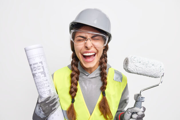 计划维护和职业概念欣喜若狂的女工程师与蓝图和穿着安全服的油漆辊合影 忙着在白色墙壁上隔离的施工现场重建眼镜头盔手套