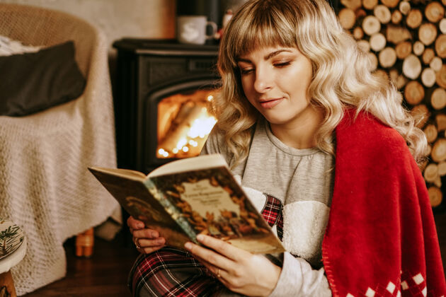女性快乐的年轻女子在圣诞室内前看书壁炉女孩穿着舒适的家常衣服 穿着温暖的红色格子呢放松房间假日