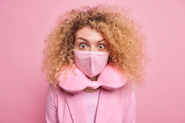 距离在冠状病毒大流行期间 一位头发卷曲浓密的欧洲妇女戴着防护面具 戴着颈枕 听到一些穿着正式服装 对着粉色墙壁摆出的令人震惊的姿势姿势女性发型