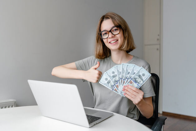 现金年轻的白种女人在家里拿着笔记本电脑 展示着金钱和拇指女性工作笔记本电脑