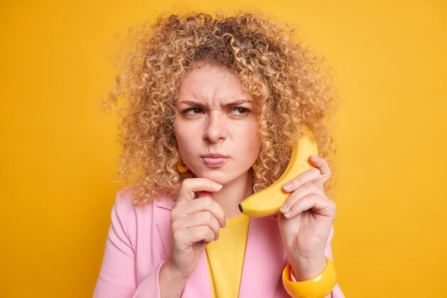 闷闷不乐严肃的卷发欧洲女人带着不满的表情假装有重要的谈话拿着美味的香蕉放在耳边打扮成正式的笑脸隔着黄色的墙壁人消极女人