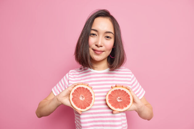 水果深色头发的漂亮黑发女人在身上抱着两个半葡萄柚 穿着休闲的条纹t恤 看起来很满足 隔着粉色的墙女人抱着多汁的水果玩自然模特