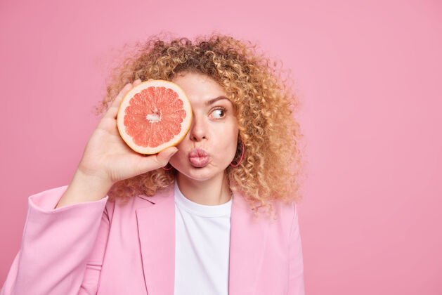 惊人健康的生活方式和维生素可爱的女人有卷曲的浓密的头发保持嘴唇折叠用一半的葡萄柚遮住眼睛保持饮食穿着正式的衣服隔离在粉红色的墙壁空白果汁素食柑橘