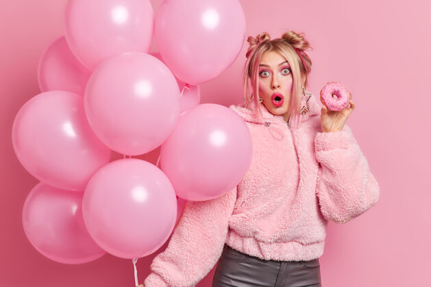 意外图为惊讶的欧洲女人 发型时髦 穿着时髦的衣服 手拿美味的甜甜圈和气球 享受生日派对 震惊地得到意想不到的礼物模型一切都是粉红色的甜点生日美味