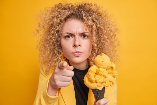发型年轻的卷发女人看起来很生气 直接指责你拿着美味的冰激凌吃着美味的夏日甜点穿着正式的衣服隔着黄色的墙人青少年美味