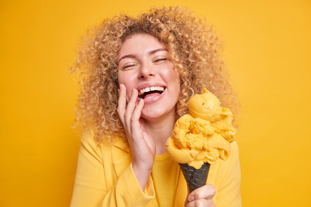 人类照片中喜出望外的卷发女人觉得很有趣微笑睁大眼睛享受吃芒果味的美味冰淇淋摆出黄色墙壁上隔离着美味的冷冻甜点年轻夹克脸