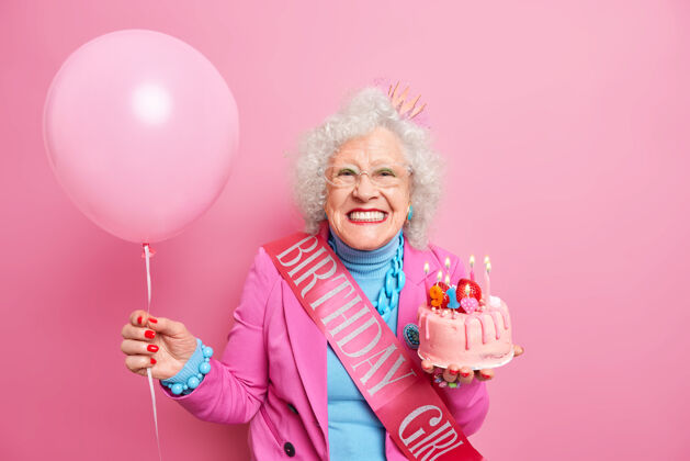 快乐快乐满脸皱纹的女退休老人的摄影棚 浓妆艳抹 笑得牙牙学语 拿着燃着蜡烛的喜庆蛋糕 带着喜庆的心情提着充气气球庆典气球灰色