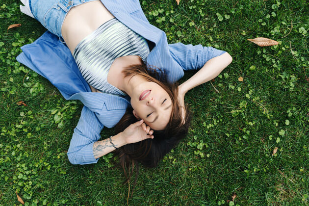 欢呼一位身着蓝色条纹服装的年轻女子的时尚肖像女士夹克躺在城市公园的绿色草坪上女人姿势女士