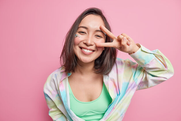 成功可爱积极的亚洲女人 深色头发 展示v字和平手势 看起来很快乐 穿着休闲衬衫 享受美好的一天 隔着粉色的墙壁 表达无忧无虑的情绪模特民族真诚