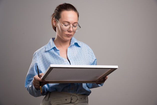 爱好戴着眼镜的女人拿着画布 在灰色上画笔高质量的照片艺术家女设备