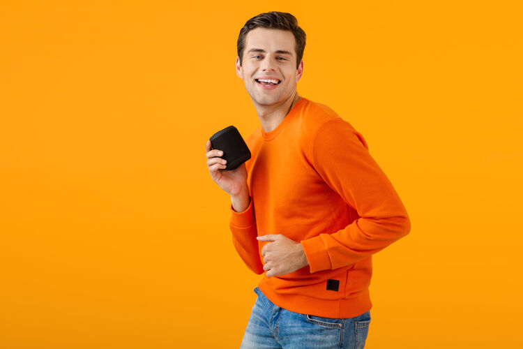 姿势时尚微笑的年轻人穿着橙色毛衣手持无线扬声器快乐地听音乐玩得开心科技搞笑积极
