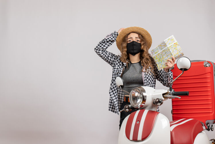 轻便摩托车正面图：戴着黑色面具的年轻旅行者女孩拿着地图站在红色轻便摩托车旁边持摩托车度假