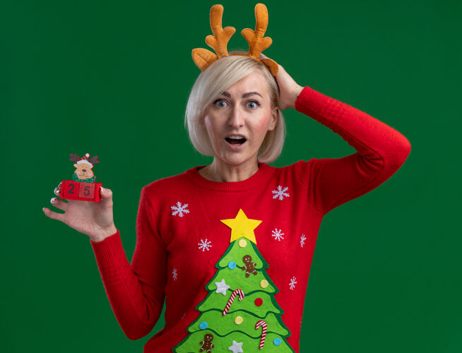 驯鹿令人印象深刻的中年金发女人戴着圣诞驯鹿鹿角头带和圣诞毛衣看着手放在头上拿着圣诞驯鹿玩具与日期隔离在绿色墙上举行手中年