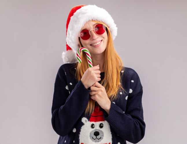 年轻人微笑的年轻漂亮女孩戴着圣诞帽戴着圣诞眼镜拿着圣诞糖果围着隔离在白色墙上的脸戴着糖果帽子