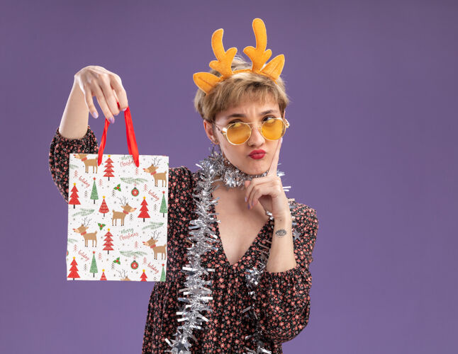 眼镜困惑的年轻漂亮女孩戴着驯鹿鹿角头带 脖子上戴着金箔花环 戴着眼镜 拿着圣诞礼品袋 看着它 触摸着隔离在紫色墙上的脸 还有复制空间漂亮困惑触摸