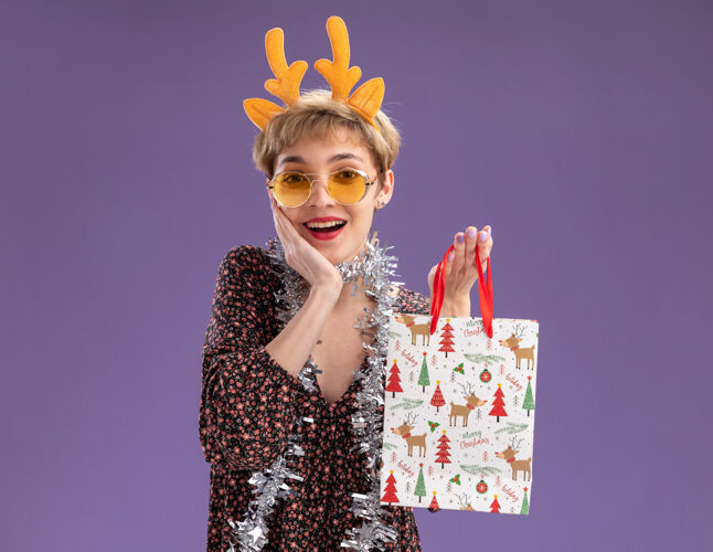 包快乐的年轻漂亮女孩戴着驯鹿鹿角头带 脖子上戴着金箔花环 戴着眼镜 手里拿着圣诞礼品袋 手放在脸上 隔离在紫色的墙上 留着复制空间头带脸女孩
