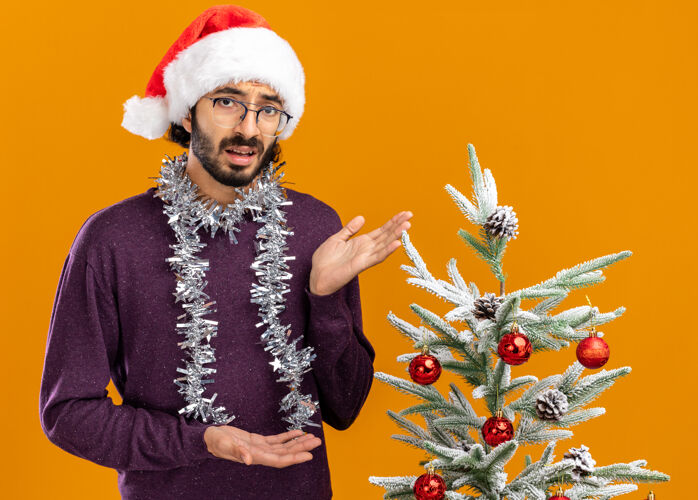 脖子站在圣诞树旁的年轻帅哥 戴着圣诞帽 脖子上戴着花环 指着隔离在橙色墙上的圣诞树点花环圣诞节