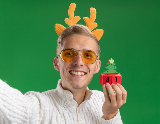 圣诞节在绿色的墙上 微笑的年轻帅哥戴着驯鹿鹿角头带戴着眼镜 手里拿着圣诞树玩具 手伸向相机 看起来很孤立年轻手家伙