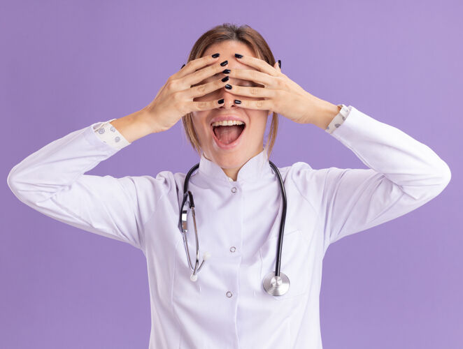穿着兴奋的年轻女医生穿着医用长袍 听诊器捂着眼睛 手被隔离在紫色的墙上人脸兴奋