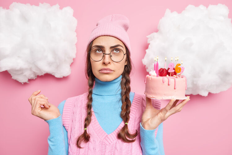 眼镜梳着两条梳辫的女孩手里拿着草莓蛋糕有着悲伤无聊的表情戴着帽子高领衫集中在粉色上面孤立着集中辫子蜡烛