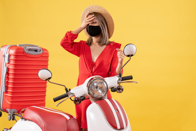 度假正面图：戴着黑色面具手持信用卡的年轻女子站在轻便摩托车红色手提箱旁流行病站着红色