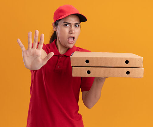 脸被吓坏了的年轻送货女孩穿着制服 戴着帽子 手里拿着披萨盒 在橙色的墙上显示着停止的手势市民人帽子