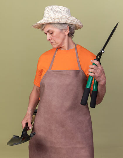 女自信的上了年纪的女园丁戴着园艺帽拿着园艺剪刀看着铲子园艺老人橄榄