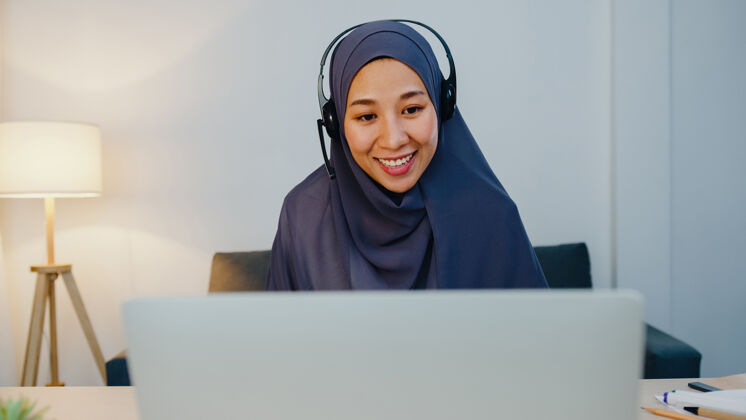通信穆斯林女士戴着耳机看网络研讨会听在线课程在晚上的家庭办公室通过电话会议进行交流迟到客户呼叫