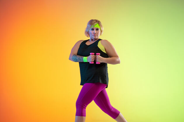 女士年轻的白人大号女模特在霓虹灯下的橙色渐变背景上训练用重量做锻炼运动的概念 健康的生活方式 积极的身体 平等模特拉伸运动