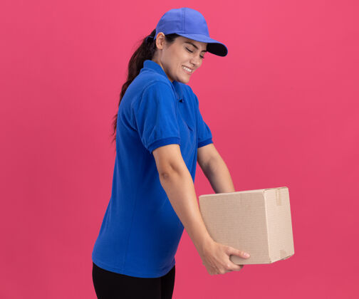 人紧张的年轻女送货员穿着制服 粉色墙上有一个装帽子的盒子帽子人穿着