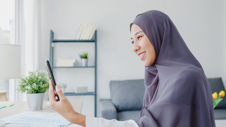 女性年轻的亚洲女商人使用智能手机通过视频聊天与朋友交谈头脑风暴在线会议 同时在客厅远程工作工作场所工作技术