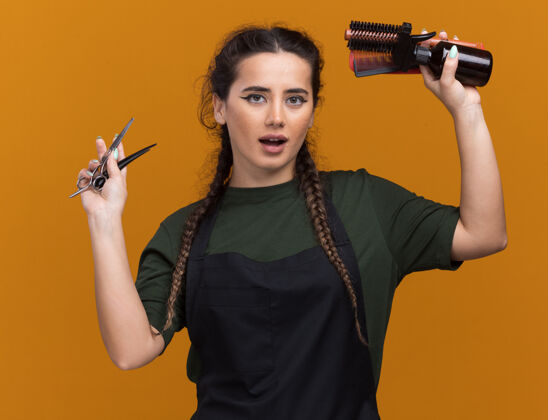 制服高兴的年轻女理发师穿着制服举起理发工具隔离在橙色的墙上衣服理发师请