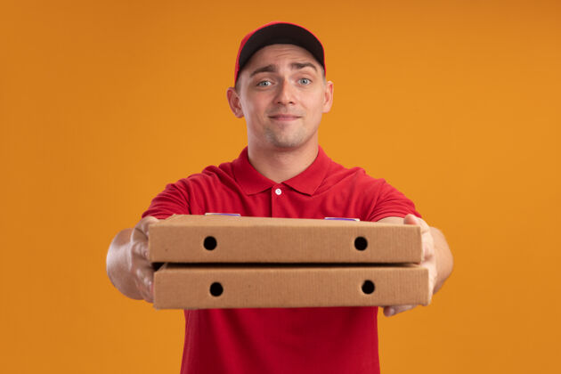 姿势年轻的送货员穿着制服 戴着帽子 拿着披萨盒 站在橘色的墙上 看着镜头盒子感觉披萨