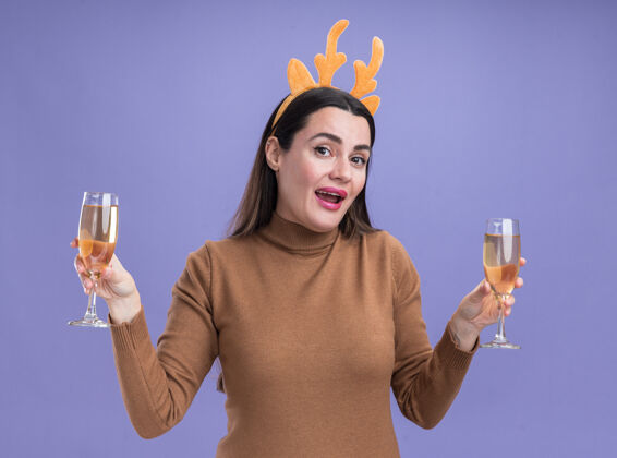 年轻微笑的年轻漂亮女孩穿着棕色毛衣 戴着圣诞发箍 手里拿着两杯香槟孤立在蓝色的墙上香槟微笑女孩