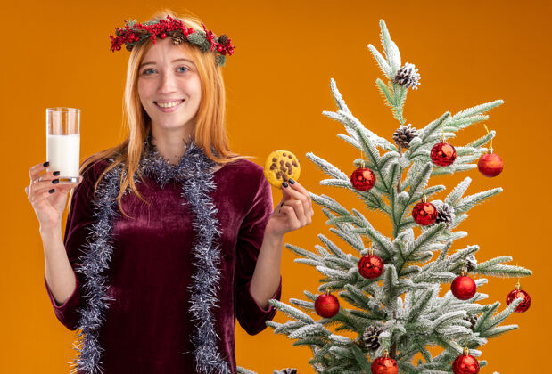 抱着站在圣诞树旁微笑的年轻漂亮女孩 身穿红色连衣裙 脖子上戴着花环 手里拿着一杯牛奶 橘黄色的墙上隔着饼干圣诞节就在附近牛奶