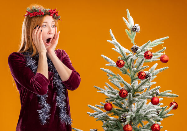 脸颊害怕的年轻漂亮女孩站在圣诞树旁边穿着红色的裙子 脖子上戴着花环看着圣诞树把双手放在脸颊上孤立在橙色的墙上花环脖子手