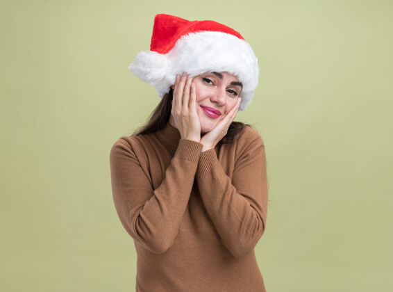 脸颊高兴的年轻漂亮的女孩戴着圣诞帽 两手孤立地贴在橄榄绿的墙上女孩橄榄穿