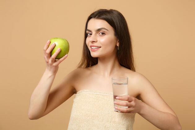 新鲜近距离拍摄完美肌肤的半裸女人 裸妆将苹果和水隔离在米色粉彩墙上手玻璃器皿水
