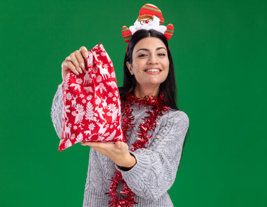 花环微笑着的年轻白人女孩戴着圣诞老人的头带 脖子上戴着金箔花环 把圣诞礼物袋伸向隔离在绿色墙上的复制空间微笑穿女孩