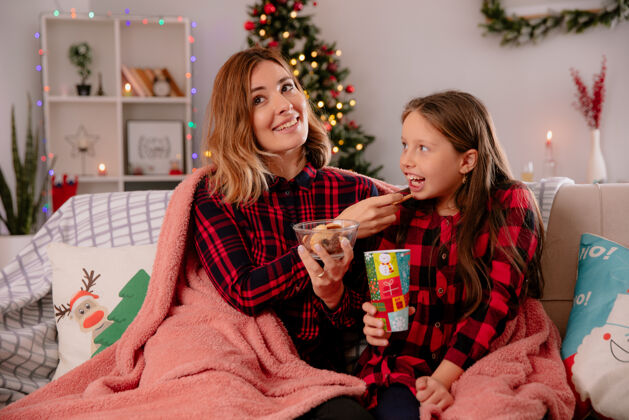 女儿高兴的母亲喂她的女儿饼干坐在沙发上覆盖着毯子和享受圣诞节的时间在家里房子坐着时间