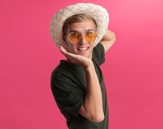 站着面带微笑的年轻帅哥 穿着绿色衬衫 戴着眼镜 戴着帽子 手放在下巴下面 隔离在粉红色的墙上人脸手